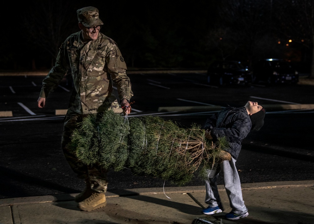 A U.S. Army soldier in OCP (Operational Camouflage Pattern) uniform and a child are carrying a bundled Christmas tree together across a parking lot at night. The soldier is smiling and holding the top end of the tree while the child, wearing a dark jacket and gray sweatpants, carries the root end. The scene appears to be part of a holiday tree distribution event, likely on or near a military installation.