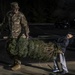 A U.S. Army soldier in OCP (Operational Camouflage Pattern) uniform and a child are carrying a bundled Christmas tree together across a parking lot at night. The soldier is smiling and holding the top end of the tree while the child, wearing a dark jacket and gray sweatpants, carries the root end. The scene appears to be part of a holiday tree distribution event, likely on or near a military installation.