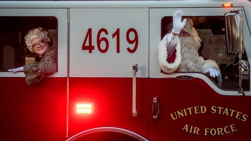 Two people dressed as Santa Claus and Mrs. Claus are riding in a United States Air Force fire truck numbered 4619. Santa is waving from the passenger window while Mrs. Claus leans out from the driver's side window, both smiling. This appears to be a holiday event or Christmas parade on a military installation.
