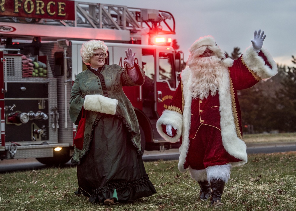 Two individuals dressed as Santa Claus and Mrs. Claus are waving to an unseen crowd in front of a red fire truck. The setting appears to be an outdoor holiday event, likely at a military or Air Force base based on the partial text visible on the fire truck. The photo is taken at dusk or early evening, giving the scene a warm, festive atmosphere.