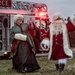 Two individuals dressed as Santa Claus and Mrs. Claus are waving to an unseen crowd in front of a red fire truck. The setting appears to be an outdoor holiday event, likely at a military or Air Force base based on the partial text visible on the fire truck. The photo is taken at dusk or early evening, giving the scene a warm, festive atmosphere.