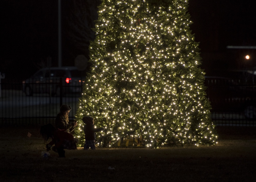 A nighttime outdoor scene featuring a large Christmas tree densely covered in warm white lights. Two figures, appearing to be an adult and a small child, are crouched near the base of the tree. The background shows a dark urban or suburban setting with a fence, parked vehicles, and distant lights.