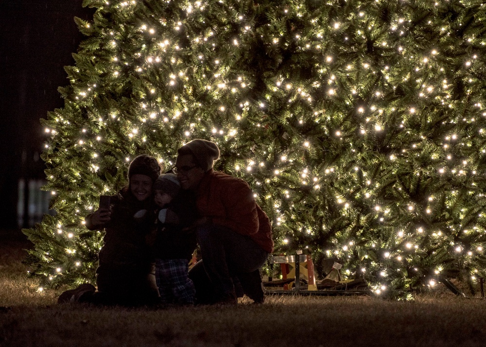 A group of three people, including at least one child, are gathered in front of a large outdoor Christmas tree illuminated with hundreds of warm white lights at night. They appear to be taking a selfie or posing for a photo together. Wrapped gifts are visible beneath the tree to the right of the group.