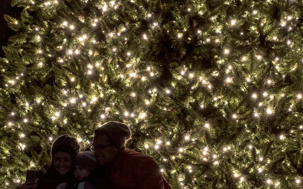 A group of three people, including at least one child, are gathered in front of a large outdoor Christmas tree illuminated with hundreds of warm white lights at night. They appear to be taking a selfie or posing for a photo together. Wrapped gifts are visible beneath the tree to the right of the group.