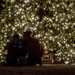 A group of three people, including at least one child, are gathered in front of a large outdoor Christmas tree illuminated with hundreds of warm white lights at night. They appear to be taking a selfie or posing for a photo together. Wrapped gifts are visible beneath the tree to the right of the group.
