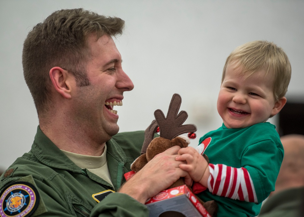 A military servicemember in an olive green flight suit is holding a young toddler, both laughing joyfully. The child is dressed in a green and red striped Christmas outfit and is holding a stuffed reindeer toy. The scene appears to be a holiday reunion or Christmas event.