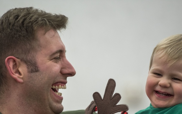 A military servicemember in an olive green flight suit is holding a young toddler, both laughing joyfully. The child is dressed in a green and red striped Christmas outfit and is holding a stuffed reindeer toy. The scene appears to be a holiday reunion or Christmas event.