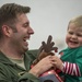 A military servicemember in an olive green flight suit is holding a young toddler, both laughing joyfully. The child is dressed in a green and red striped Christmas outfit and is holding a stuffed reindeer toy. The scene appears to be a holiday reunion or Christmas event.