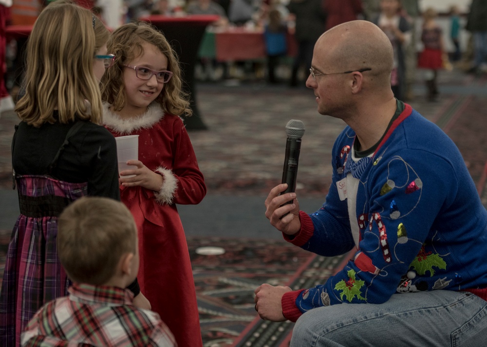 A bald adult man wearing a festive 'ugly Christmas sweater' kneels down to interview a young girl in a red Santa-style dress using a handheld microphone at what appears to be a Christmas event or party. The girl, wearing purple glasses, holds a plastic cup and smiles at the man. Two other children are visible in the foreground and left side of the frame.