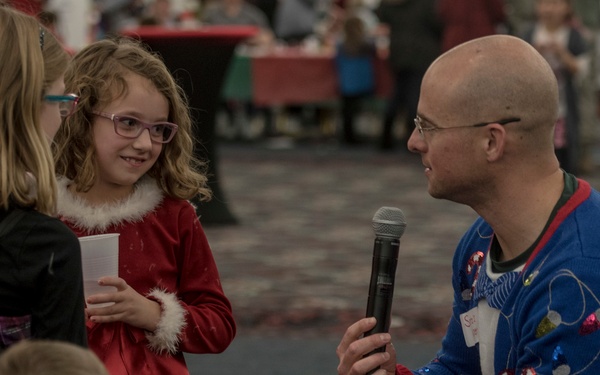 A bald adult man wearing a festive 'ugly Christmas sweater' kneels down to interview a young girl in a red Santa-style dress using a handheld microphone at what appears to be a Christmas event or party. The girl, wearing purple glasses, holds a plastic cup and smiles at the man. Two other children are visible in the foreground and left side of the frame.