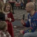 A bald adult man wearing a festive 'ugly Christmas sweater' kneels down to interview a young girl in a red Santa-style dress using a handheld microphone at what appears to be a Christmas event or party. The girl, wearing purple glasses, holds a plastic cup and smiles at the man. Two other children are visible in the foreground and left side of the frame.