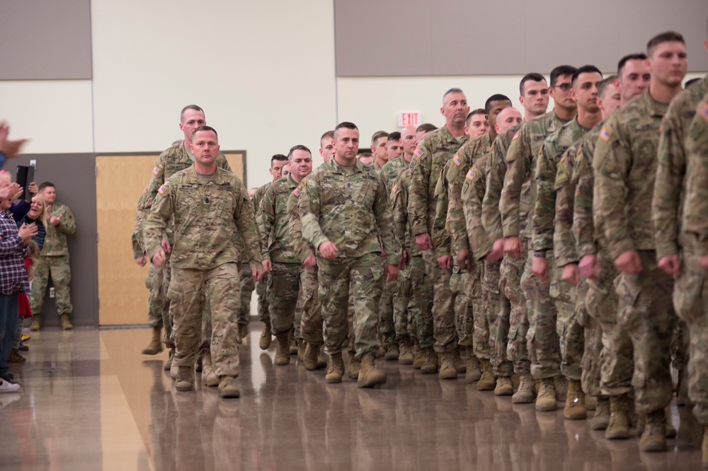A large formation of U.S. Army soldiers in OCP (Operational Camouflage Pattern) uniforms are assembled inside what appears to be a gymnasium or armory facility. Two soldiers in the foreground appear to be leading or inspecting the formation while rows of soldiers stand at attention to the right. Civilians, likely family members, are visible on the left side applauding, suggesting this may be a deployment ceremony or homecoming event.