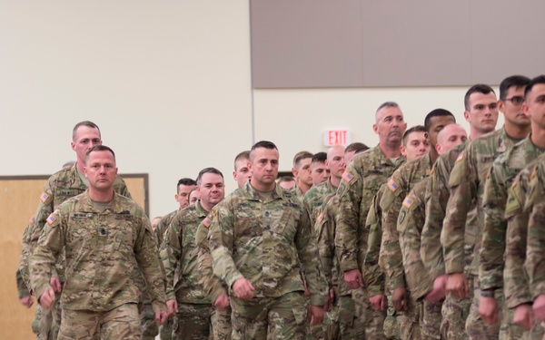 A large formation of U.S. Army soldiers in OCP (Operational Camouflage Pattern) uniforms are assembled inside what appears to be a gymnasium or armory facility. Two soldiers in the foreground appear to be leading or inspecting the formation while rows of soldiers stand at attention to the right. Civilians, likely family members, are visible on the left side applauding, suggesting this may be a deployment ceremony or homecoming event.
