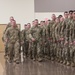 A large formation of U.S. Army soldiers in OCP (Operational Camouflage Pattern) uniforms are assembled inside what appears to be a gymnasium or armory facility. Two soldiers in the foreground appear to be leading or inspecting the formation while rows of soldiers stand at attention to the right. Civilians, likely family members, are visible on the left side applauding, suggesting this may be a deployment ceremony or homecoming event.