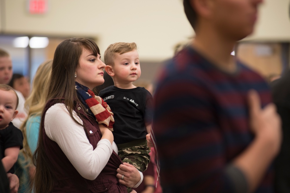 A young woman holds a toddler boy in an indoor gathering with multiple people present. The child is wearing a black shirt with 'U.S. ARMY' text and camouflage pants, while the woman wears an American flag scarf. The setting appears to be a military-related family event or homecoming ceremony.