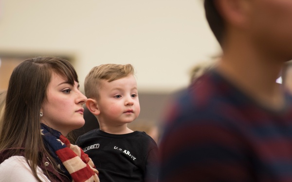 A young woman holds a toddler boy in an indoor gathering with multiple people present. The child is wearing a black shirt with 'U.S. ARMY' text and camouflage pants, while the woman wears an American flag scarf. The setting appears to be a military-related family event or homecoming ceremony.