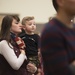 A young woman holds a toddler boy in an indoor gathering with multiple people present. The child is wearing a black shirt with 'U.S. ARMY' text and camouflage pants, while the woman wears an American flag scarf. The setting appears to be a military-related family event or homecoming ceremony.