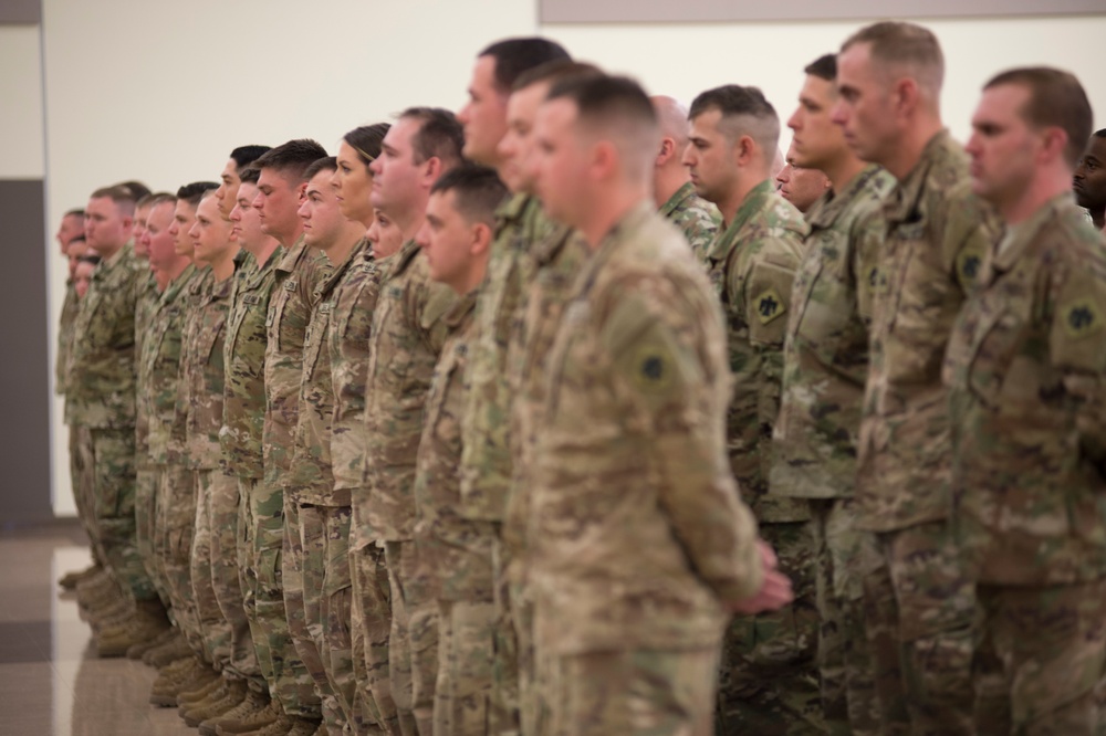 A large formation of military personnel dressed in MultiCam/OCP camouflage uniforms are standing at attention in an indoor facility. The soldiers appear to be participating in a formal military ceremony or formation event. The group includes both male and female service members arranged in multiple rows.