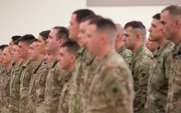 A large formation of military personnel dressed in MultiCam/OCP camouflage uniforms are standing at attention in an indoor facility. The soldiers appear to be participating in a formal military ceremony or formation event. The group includes both male and female service members arranged in multiple rows.