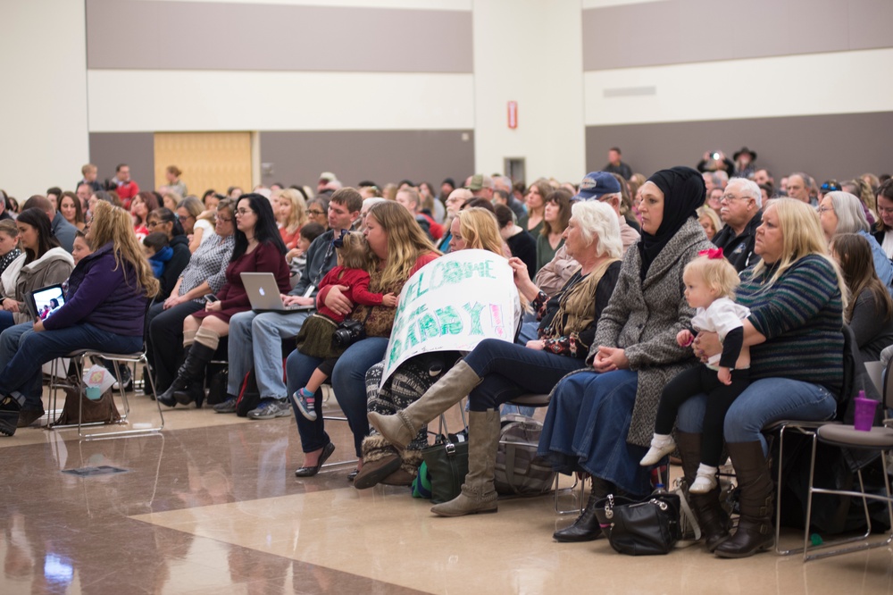 A large crowd of people is seated in rows of chairs inside what appears to be a gymnasium or large community hall. A woman in the foreground is holding a handmade sign that reads 'WELCOME BABY!!' suggesting this may be a homecoming or welcome event. The audience appears to be waiting for or watching a presentation or ceremony.