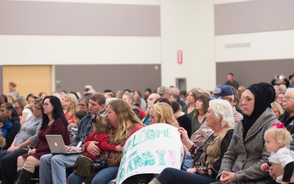A large crowd of people is seated in rows of chairs inside what appears to be a gymnasium or large community hall. A woman in the foreground is holding a handmade sign that reads 'WELCOME BABY!!' suggesting this may be a homecoming or welcome event. The audience appears to be waiting for or watching a presentation or ceremony.