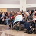 A large crowd of people is seated in rows of chairs inside what appears to be a gymnasium or large community hall. A woman in the foreground is holding a handmade sign that reads 'WELCOME BABY!!' suggesting this may be a homecoming or welcome event. The audience appears to be waiting for or watching a presentation or ceremony.