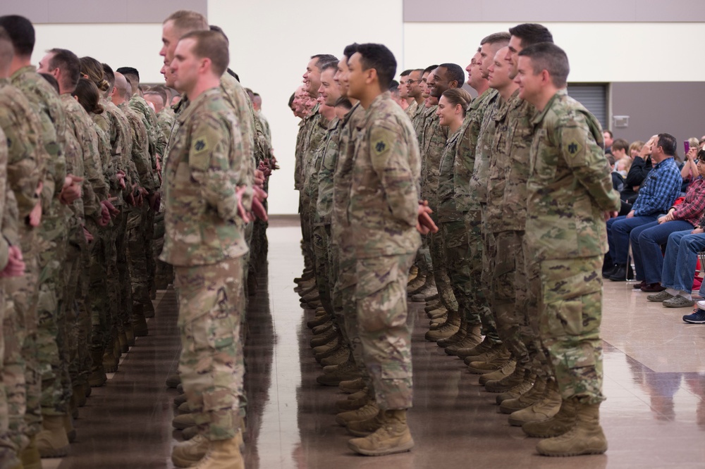 A large formation of U.S. Army soldiers in OCP (Operational Camouflage Pattern) uniforms are standing at attention or parade rest inside an indoor facility. The event appears to be a formal military ceremony, likely a deployment send-off or welcome home event, as civilian family members are seated and observing in the background. The soldiers are arranged in multiple rows facing the same direction.