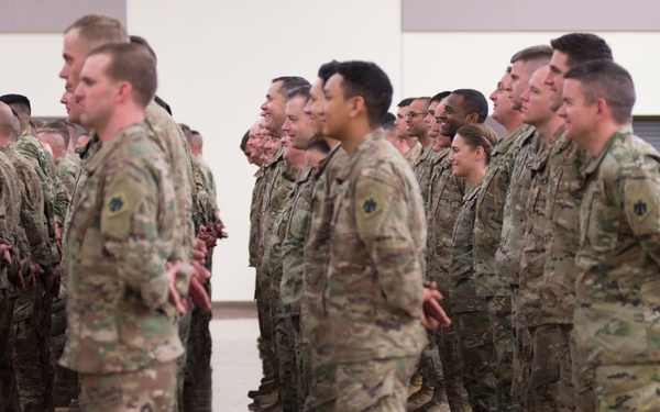 A large formation of U.S. Army soldiers in OCP (Operational Camouflage Pattern) uniforms are standing at attention or parade rest inside an indoor facility. The event appears to be a formal military ceremony, likely a deployment send-off or welcome home event, as civilian family members are seated and observing in the background. The soldiers are arranged in multiple rows facing the same direction.