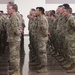 A large formation of U.S. Army soldiers in OCP (Operational Camouflage Pattern) uniforms are standing at attention or parade rest inside an indoor facility. The event appears to be a formal military ceremony, likely a deployment send-off or welcome home event, as civilian family members are seated and observing in the background. The soldiers are arranged in multiple rows facing the same direction.
