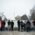 First Snow of the Season at Arlington National Cemetery A group of visitors stands in the snow at what appears to be the Eternal Flame at Arlington National Cemetery, with the Arlington House (Robert E. Lee Memorial) visible in the background on a hill. A flag is flying at half-staff, suggesting a period of national mourning. The scene is overcast and snowy, creating a somber, wintry atmosphere.