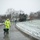 First Snow of the Season at Arlington National Cemetery A grounds worker in a high-visibility yellow safety jacket pushes a green Scotts broadcast spreader along a curved stone pathway during a light snowfall. The scene appears to be a public park or monument grounds, with snow lightly dusting the grass and trees. A few other pedestrians are visible in the background along the path.
