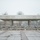 First Snow of the Season at Arlington National Cemetery A brutalist-style concrete canopy structure with cylindrical columns stands in a public space during winter snowfall. The structure is flanked by a low stone retaining wall with a row of blue/purple flowering plants, likely pansies, despite the snowy conditions. The foreground features a wet granite or stone tile plaza, and bare deciduous trees are visible in the background under an overcast sky.