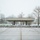 First Snow of the Season at Arlington National Cemetery A brutalist-style concrete pavilion or shelter structure stands in an open park or campus setting during winter conditions with light snow dusting. The scene is stark and deserted, with bare deciduous trees and snow-covered ground vegetation surrounding the flat-roofed concrete structure. The overcast sky and muted tones create a cold, quiet atmosphere.