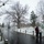 First Snow of the Season at Arlington National Cemetery A snowy winter scene at what appears to be a cemetery or memorial park, with a wet winding road flanked by chain-link bollard barriers. Two visitors stand in the foreground examining an informational map or sign, while a small group of people walks along the road in the background. Bare deciduous trees and light snow cover the grounds, creating a somber, overcast atmosphere.