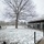 First Snow of the Season at Arlington National Cemetery A winter scene at what appears to be a public park or historic site, with a light dusting of snow covering the ground and grass. A large bare deciduous tree dominates the center of the image, flanked by a covered pergola or colonnade structure on the right where a trolley bus is parked. Several visitors are visible walking along pathways in cold-weather clothing.