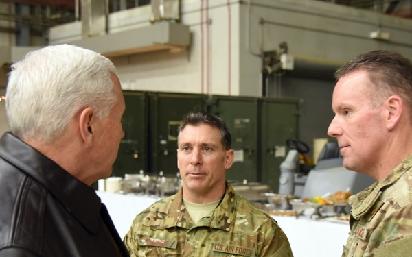 A civilian in a leather jacket with an American flag patch is engaged in conversation with two U.S. Air Force personnel in OCP (Operational Camouflage Pattern) uniforms inside what appears to be a military hangar or facility. One of the military personnel displays a brigadier general star insignia and 'U.S. AIR FORCE' tape visible on their uniform. A buffet-style food setup is visible in the background, suggesting a troop visit or morale event.