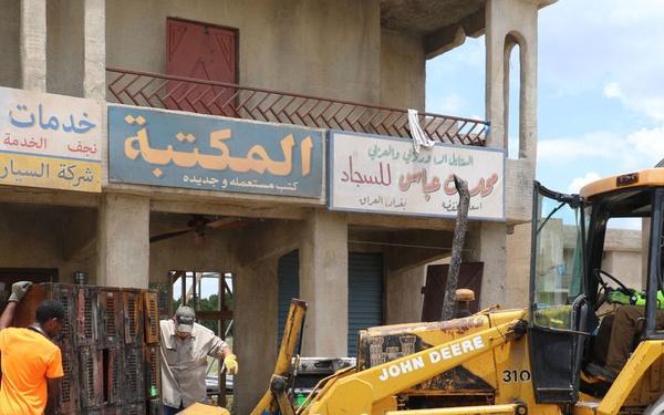 A John Deere 310 backhoe loader is being operated in front of a row of commercial shops with Arabic signage. Several workers are present, handling materials and debris in what appears to be a construction, demolition, or cleanup operation. The scene takes place in an urban area with storefronts visible in the background.