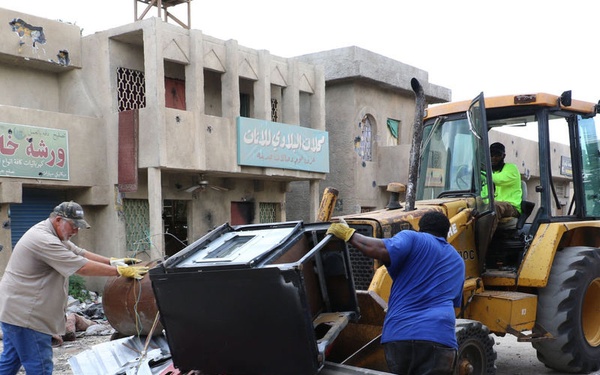 Workers are clearing debris and damaged materials from a street using a yellow front-loader/bulldozer in what appears to be an urban area with Arabic-language signage. Two workers on the ground are handling twisted metal and damaged equipment while a third operates the heavy machinery. The surrounding buildings show visible damage including bullet holes and general deterioration, suggesting post-conflict cleanup operations.