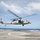 A military helicopter, consistent with an MH-60 Seahawk variant, is hovering just above a naval vessel's flight deck over open ocean. The side door is open with at least one crew member visible inside. The rotors are in motion, indicating active flight operations.