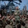 A senior military officer in U.S. Marine Corps MARPAT camouflage uniform addresses a large group of seated Marines on what appears to be the flight deck of a naval vessel. The speaker stands at the center while dozens of Marines sit around him in a semicircle, attentively listening. Military helicopters, including what appears to be a CH-53 or similar heavy-lift helicopter, are visible in the background.