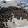 A large formation of military personnel, likely U.S. Marines and Navy sailors, are assembled on the flight deck of an amphibious assault ship at sea. Several military helicopters, including what appear to be UH-1Y Venom and UH-60 variants, are parked on the deck's edges. One or two individuals appear to be addressing the assembled group, suggesting a briefing or all-hands meeting.
