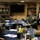 A group of military personnel, primarily in U.S. Marine Corps camouflage uniforms, are seated around a blue-covered table in what appears to be a briefing or conference room aboard a naval vessel. One individual in the center appears to be actively speaking and gesturing, suggesting a leadership briefing or planning session. The room contains bookshelves, a wall-mounted television, and various items on the table consistent with a working meeting.