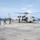 A military helicopter, identified by the number '41' on its fuselage, is on the flight deck of a naval vessel at sea. Several personnel in flight suits and military gear are approaching or attending to the helicopter, which has its rotors spinning. The scene depicts active flight deck operations with the open ocean visible in the background.
