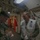 Two U.S. Marines are interacting inside what appears to be a naval vessel corridor or passageway. One Marine in camouflage uniform is writing in a notepad while the other, wearing a white safety vest and holding a helmet, is smiling. The setting features overhead cable runs and bulkhead-style architecture typical of a military ship interior.