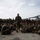 A military officer in camouflage uniform walks through a large group of seated and standing soldiers on what appears to be the flight deck of a naval vessel. The central figure appears to be addressing or briefing the assembled troops, who are gathered in a semicircle around him. Military helicopters are visible in the background, confirming the naval ship flight deck setting.