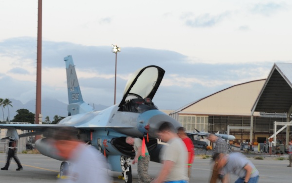 A military flight line scene showing ground crew personnel conducting post-flight or pre-flight operations around an F-16 Fighting Falcon jet fighter on a tarmac. The aircraft's canopy is open, suggesting a pilot may be exiting or the aircraft is being prepared. The setting appears to be a tropical military air base, likely in Hawaii, based on the palm trees and mountain backdrop.