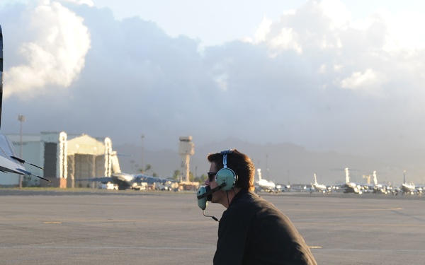 A ground crew technician is crouching on an airport tarmac near the tail section of an aircraft, wearing aviation headphones with a microphone and connected by a communication cable. The scene appears to be at dusk or dawn based on the warm, low-angle lighting. Multiple aircraft and airport infrastructure, including hangars and a control tower, are visible in the background.