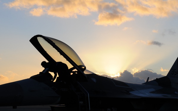 A silhouetted F-16 Fighting Falcon fighter jet is parked on a flight line tarmac at sunset or sunrise. The tail markings 'AK' and '35 FW' indicate the aircraft belongs to the 35th Fighter Wing based at Misawa Air Base, Alaska/Japan. A person, likely a pilot or crew chief, is visible climbing into or out of the cockpit.
