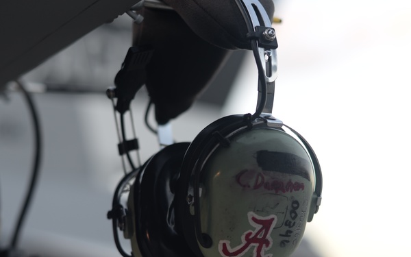 A pair of aviation headphones is hanging from what appears to be an aircraft structure. The headset has a University of Alabama 'A' logo sticker and handwritten text on the ear cup. The background is blurred, showing an outdoor airfield or tarmac environment.