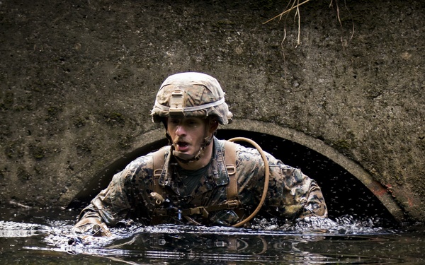 A military service member in full combat gear is emerging from a concrete culvert tunnel through water, likely during a training exercise. The soldier is wearing camouflage utilities, a combat helmet, and tactical vest with a hydration pack. The still water in the foreground creates a clear mirror reflection of the soldier and the arched tunnel opening.